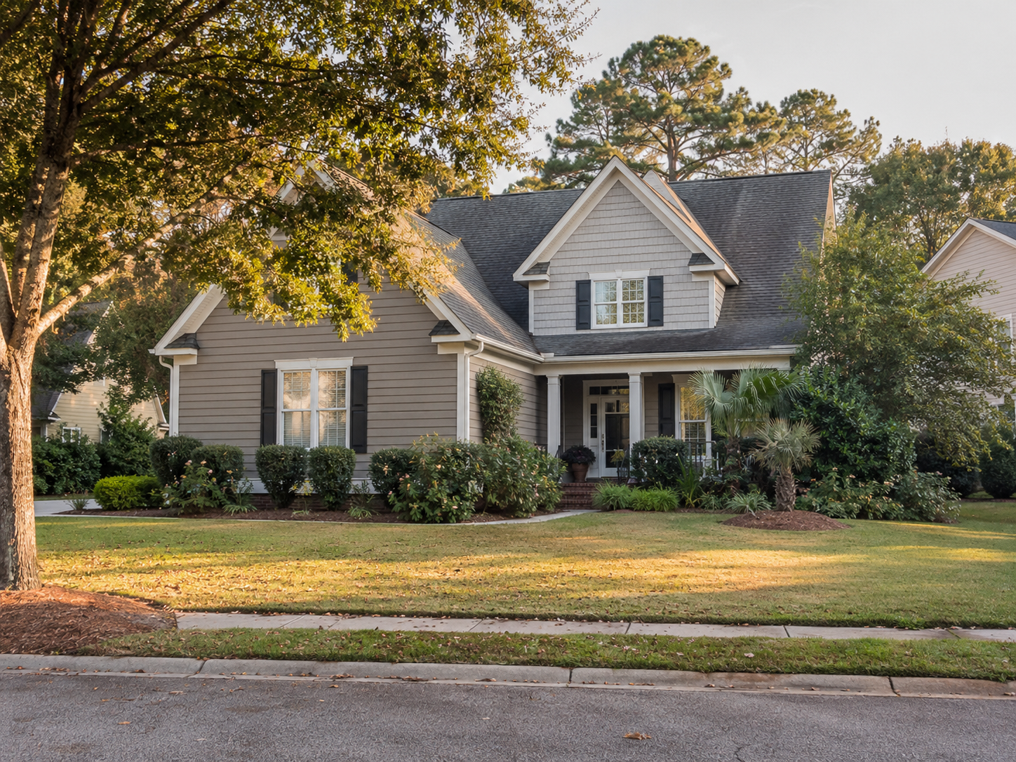 Two-story home with tan siding, black shutters, a covered front porch, and a manicured lawn on a tree-lined street in late afternoon light.